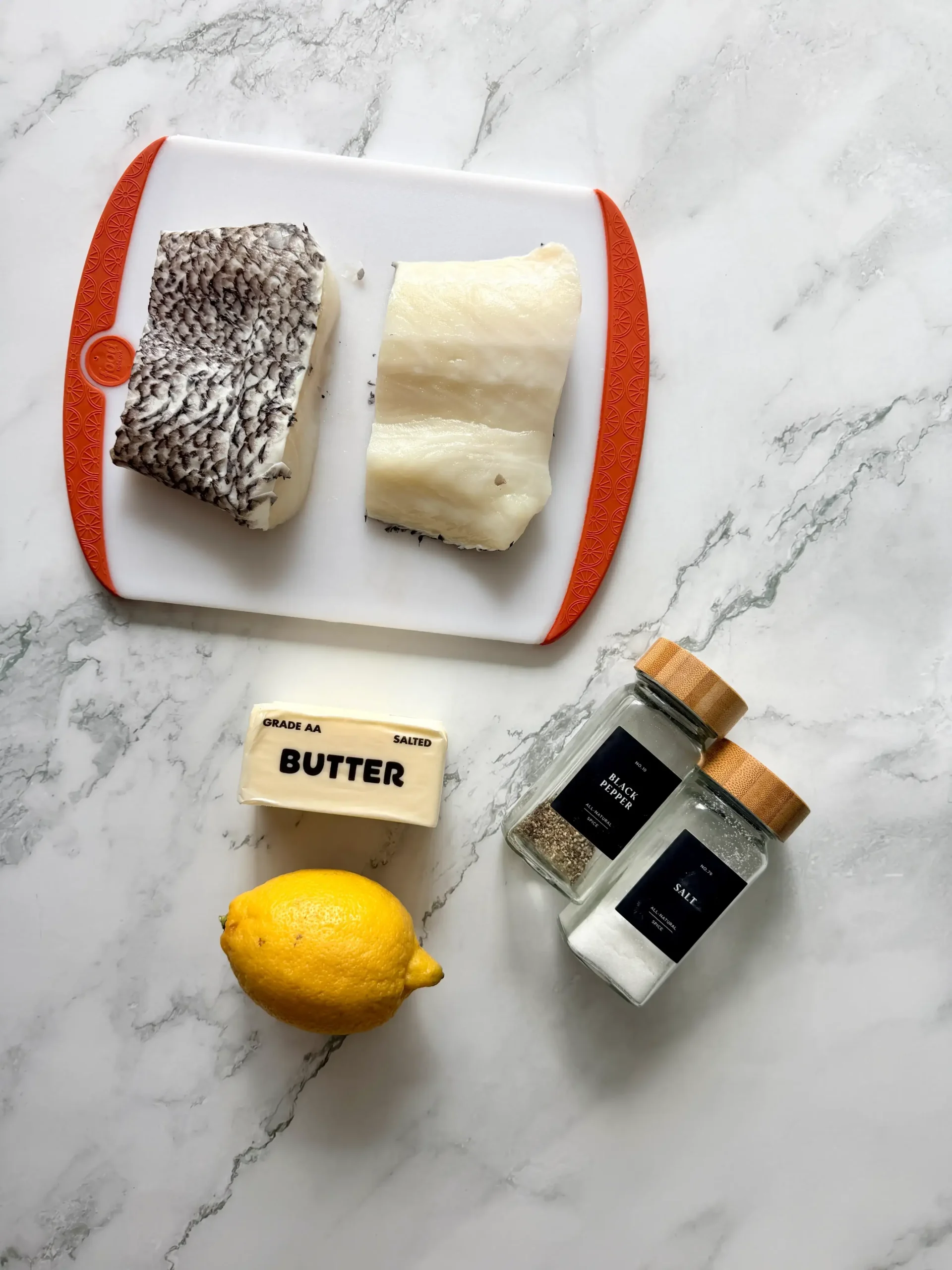 Two raw Chilean sea bass fillets on a white cutting board with butter, lemon, salt, and black pepper on a marble countertop.