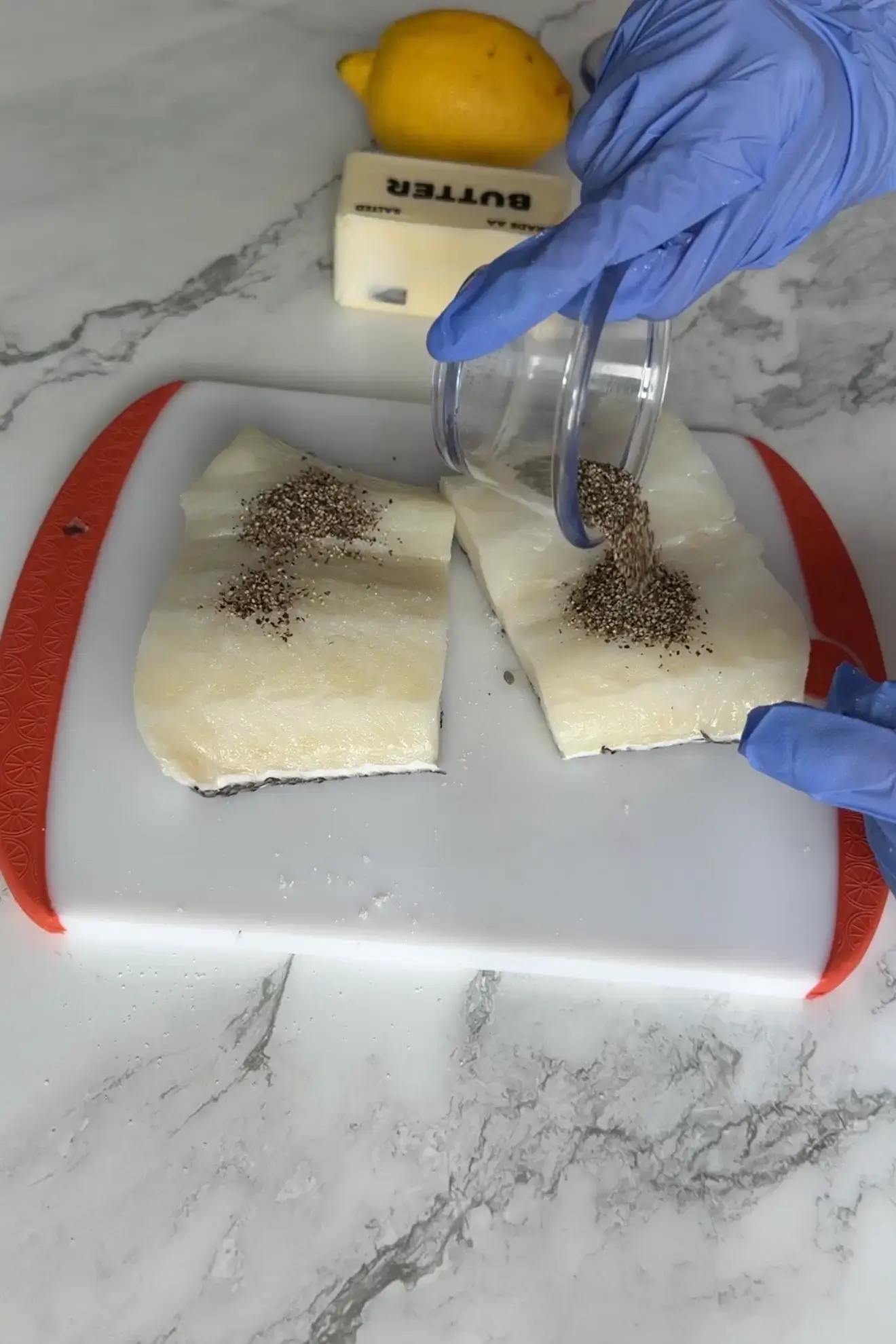 Fresh sea bass fillets on a cutting board as ground black pepper is sprinkled over the flesh; butter and a lemon sit in the background.