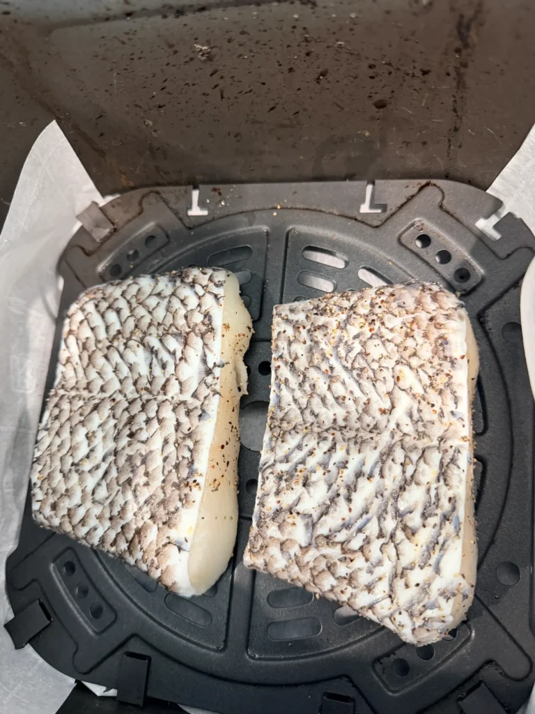 Two seasoned sea bass fillets placed skin-side up in an air fryer basket lined with parchment paper.