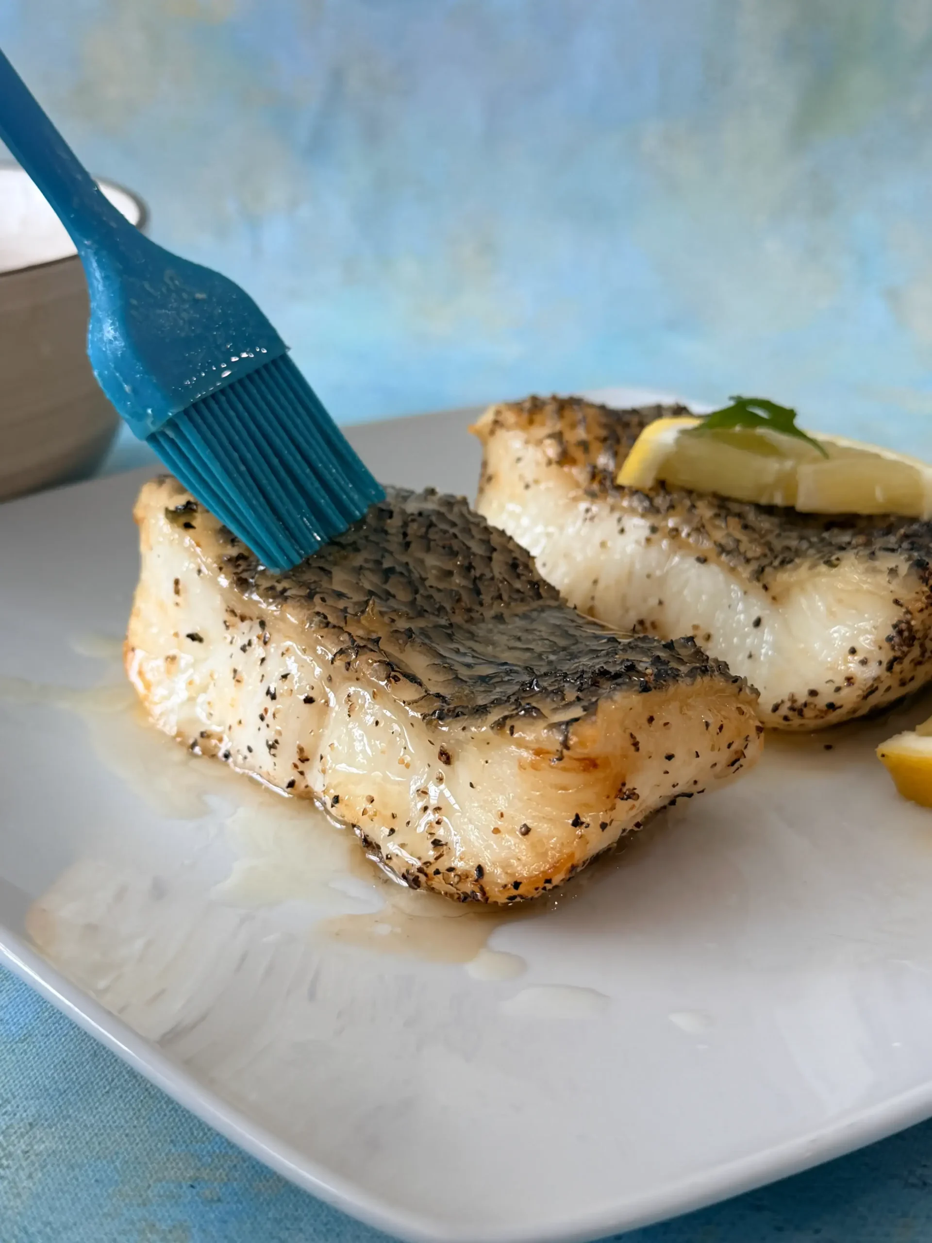 Cooked sea bass fillets on a white plate while a blue silicone brush spreads melted lemon butter over the top.