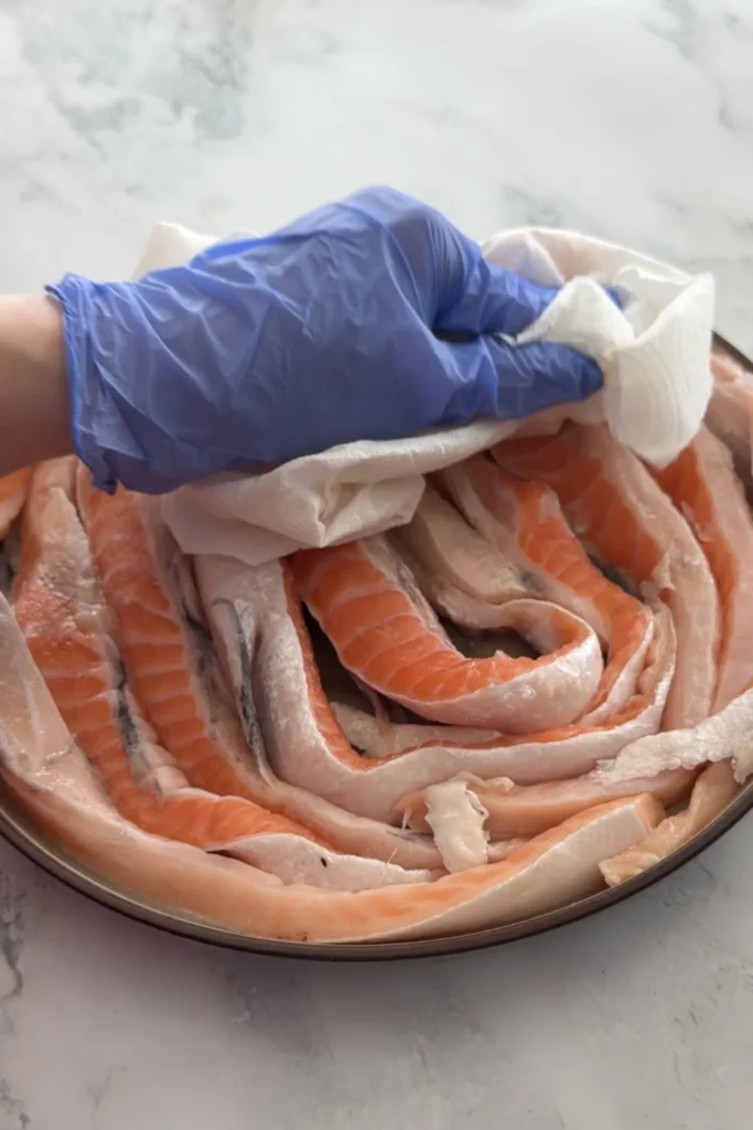 Hand patting dry raw salmon belly strips with a paper towel.