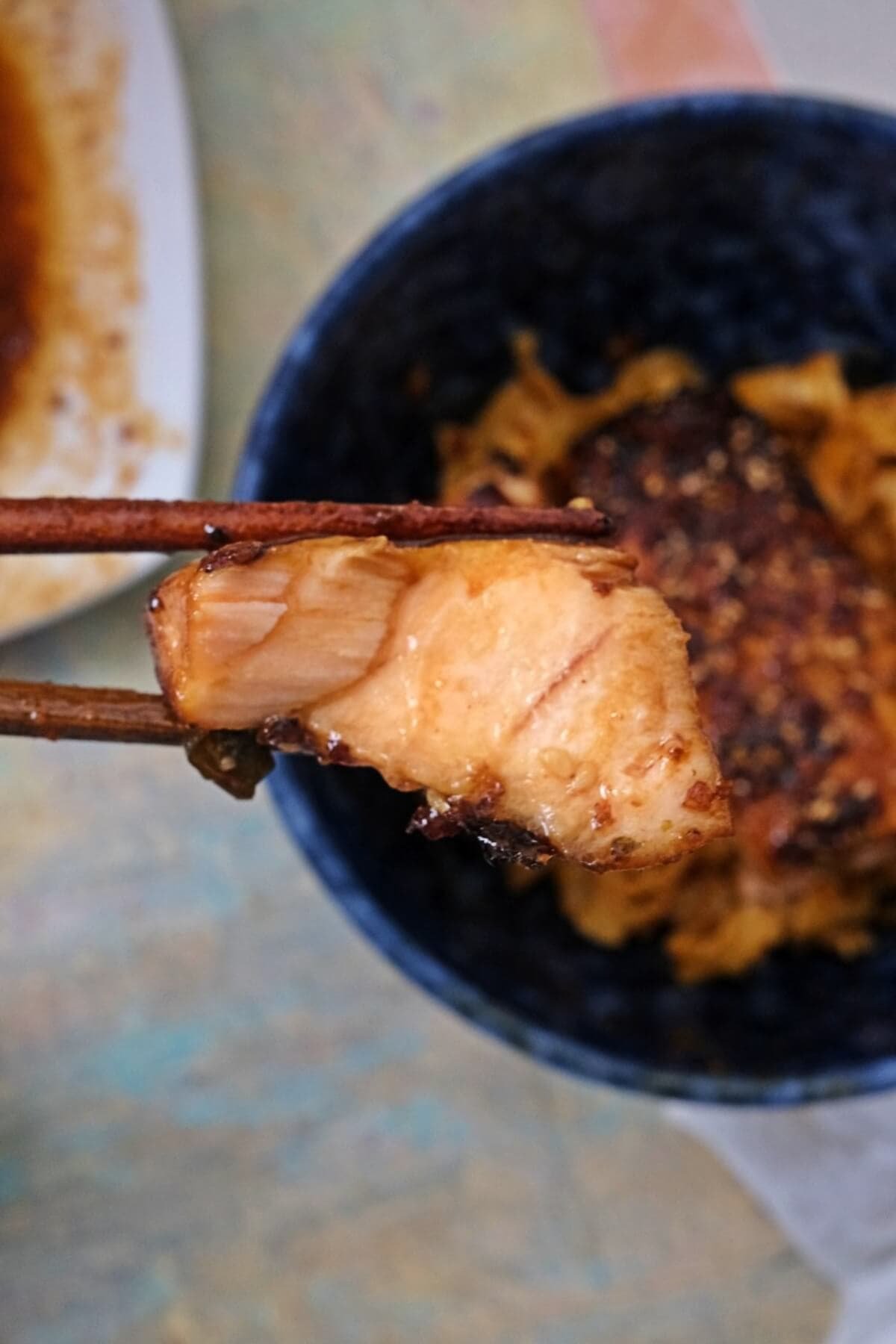 Air fryer teriyaki salmon held with chopsticks, showing a juicy, glazed interior with a lightly caramelized exterior, served over rice in a bowl.