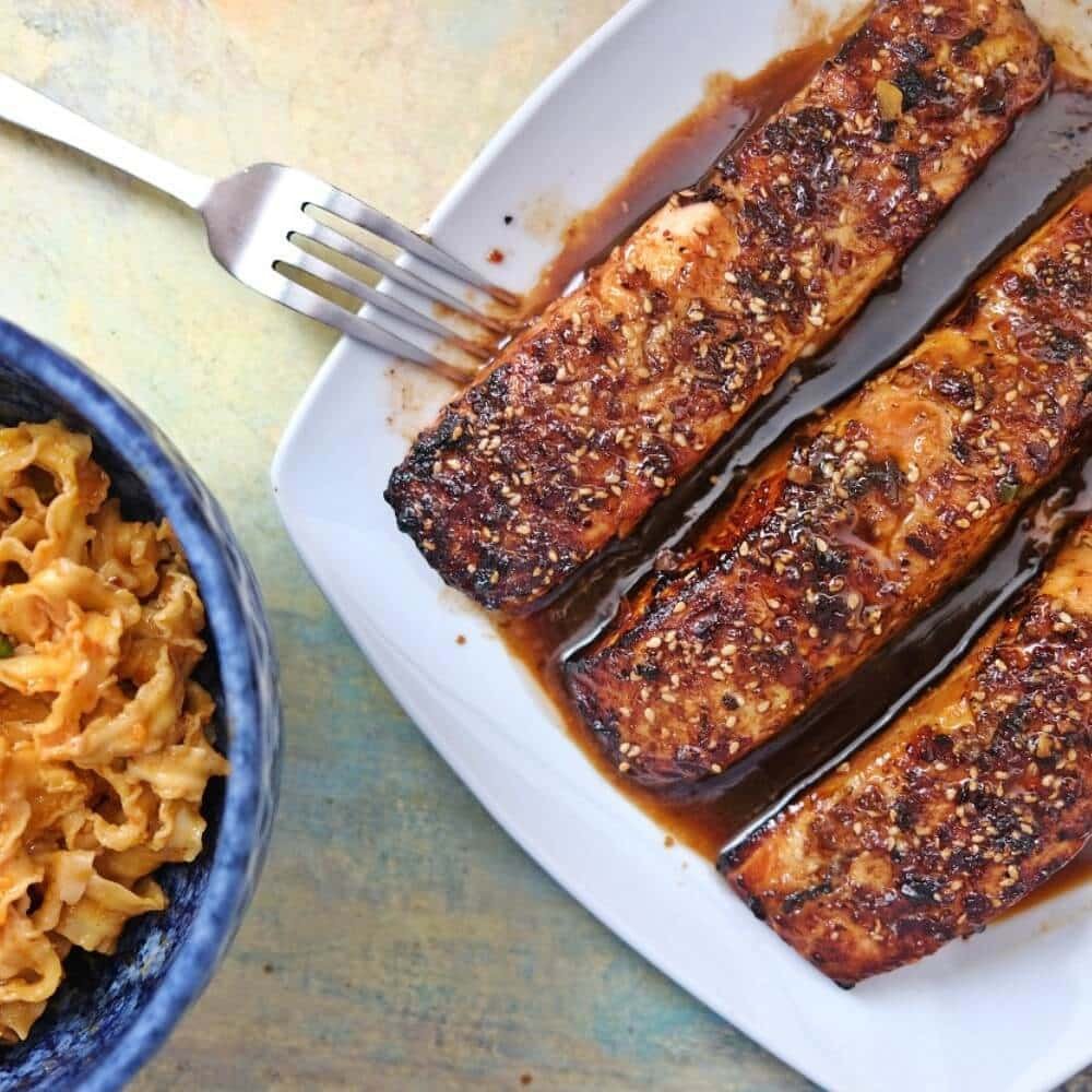 Air fryer teriyaki salmon fillets glazed with sauce and sprinkled with sesame seeds on a white plate, served with a side of noodles.