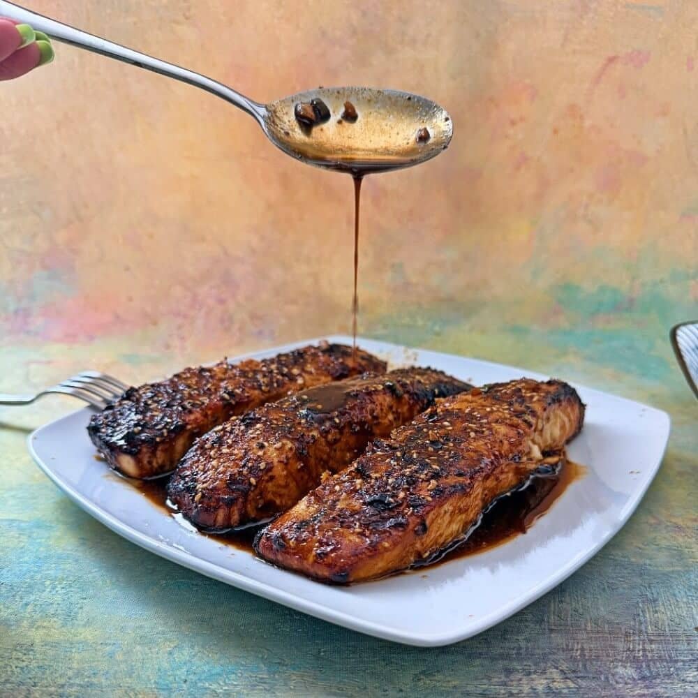 Air fryer teriyaki salmon fillets on a white plate, topped with sesame seeds and drizzled with glossy teriyaki sauce from a spoon above.