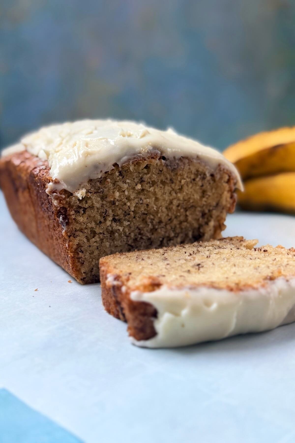 Banana bread loaf topped with cream cheese frosting, sliced on a white surface with ripe bananas in the background