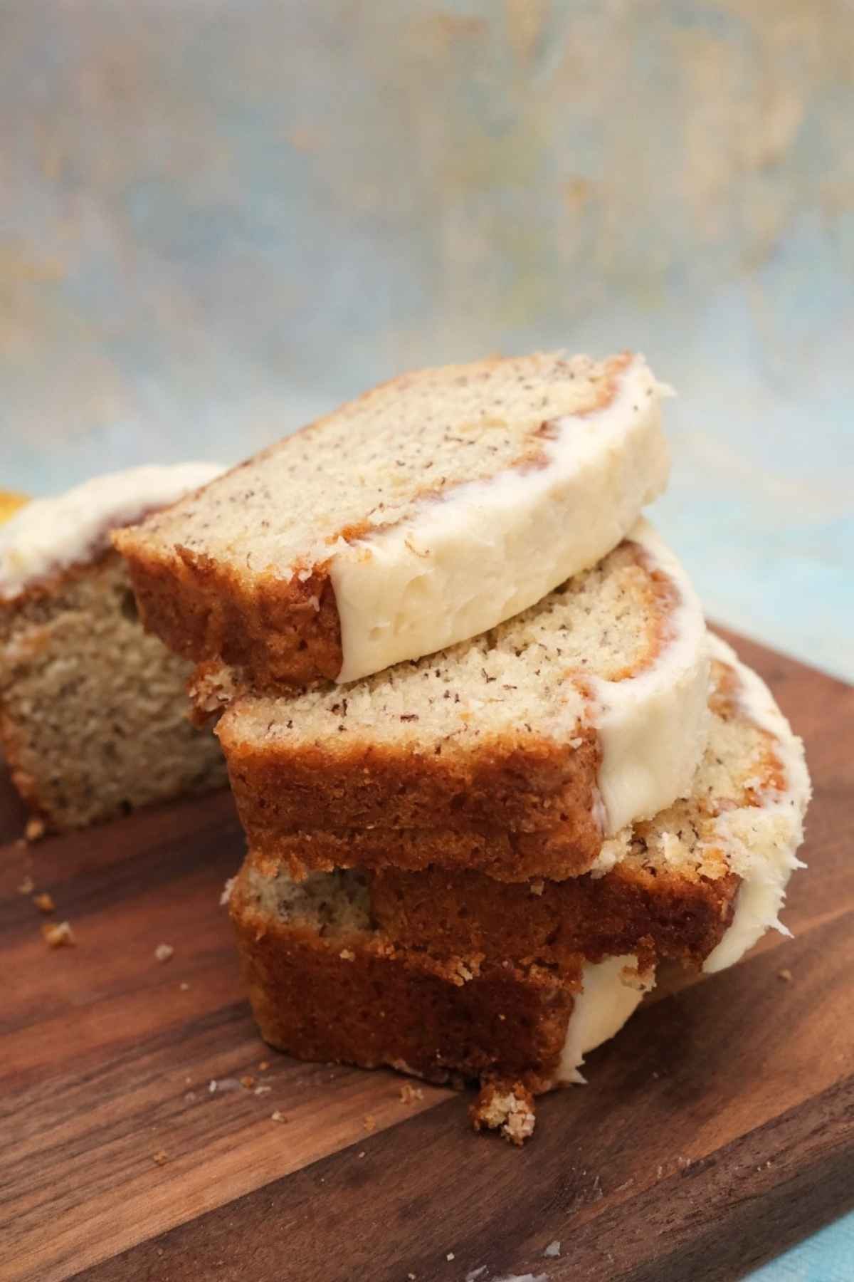 Stacked slices of banana bread with cream cheese frosting on a wooden board with a knife and ripe bananas nearby.