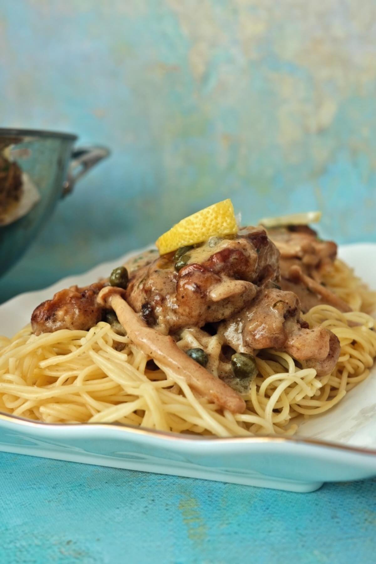 Creamy chicken piccata served over angel hair pasta with lemon wedge and capers on a white plate.