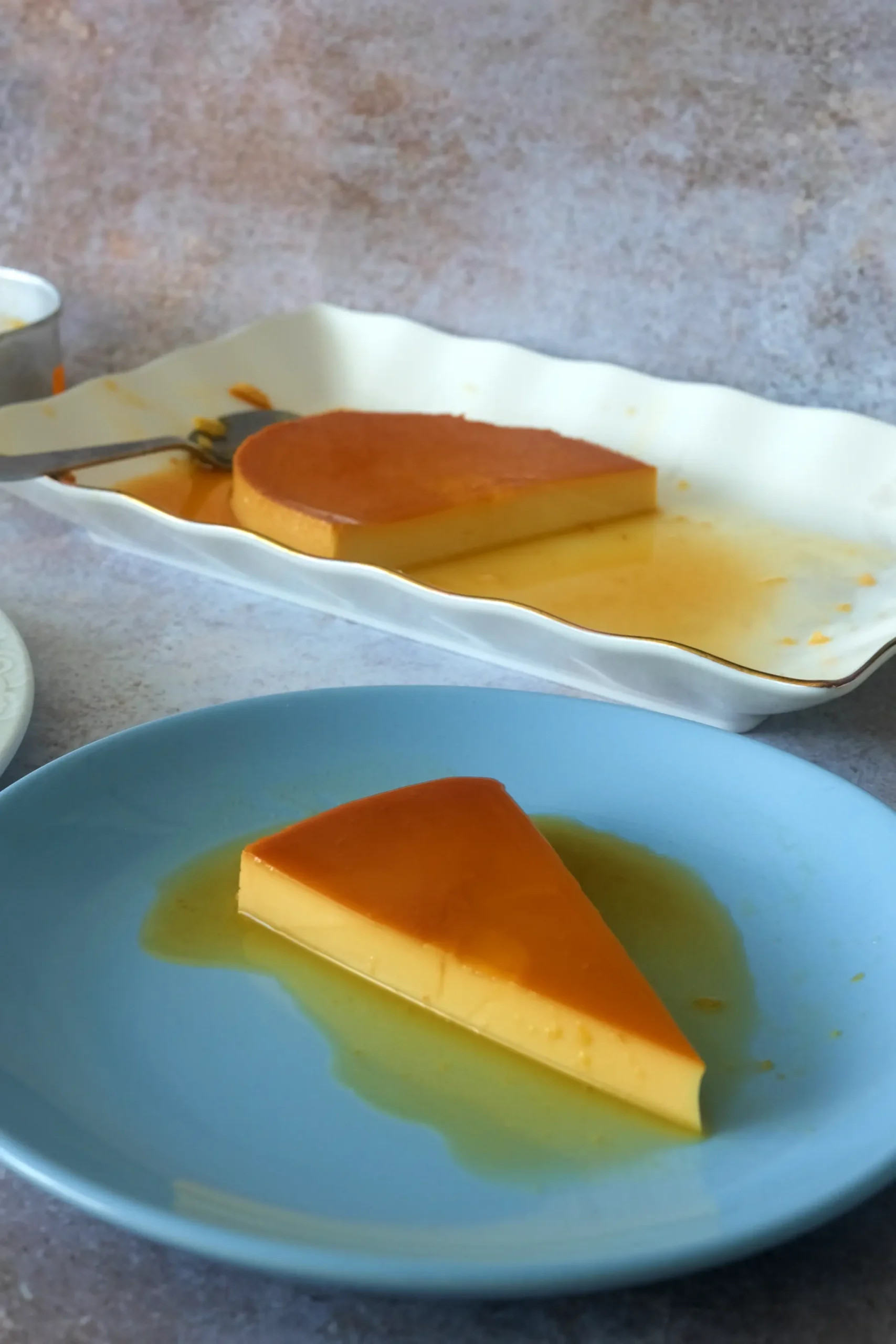 Slice of leche flan in the foreground with the remaining flan on a white platter in the background