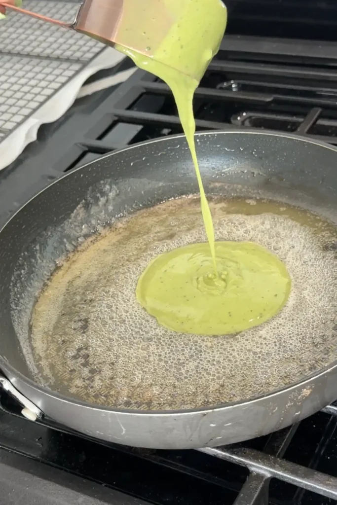 Pouring green matcha pancake batter from measuring cup into buttered skillet on stovetop.