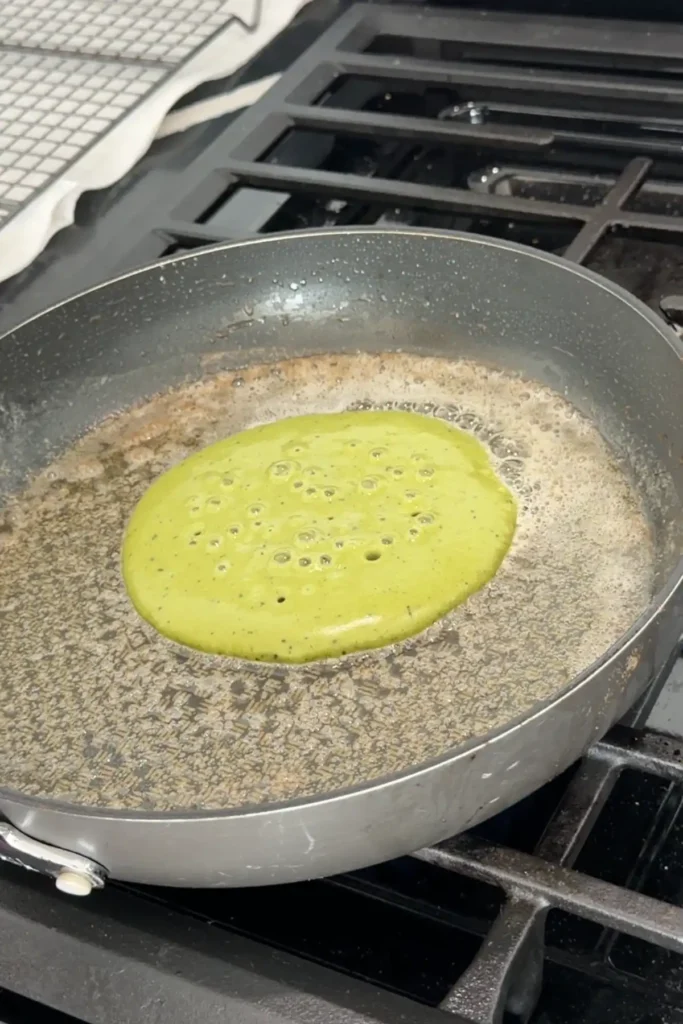 Matcha pancake cooking in skillet with bubbles forming on the surface before flipping.