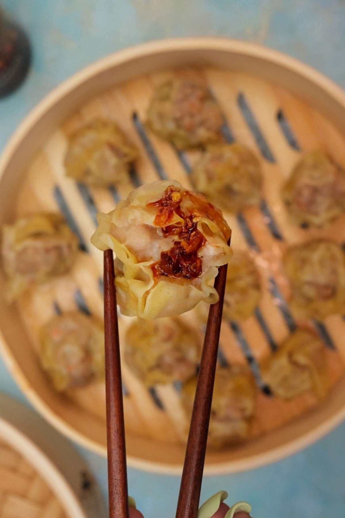 Pork siomai topped with chili garlic held with chopsticks over a bamboo steamer.