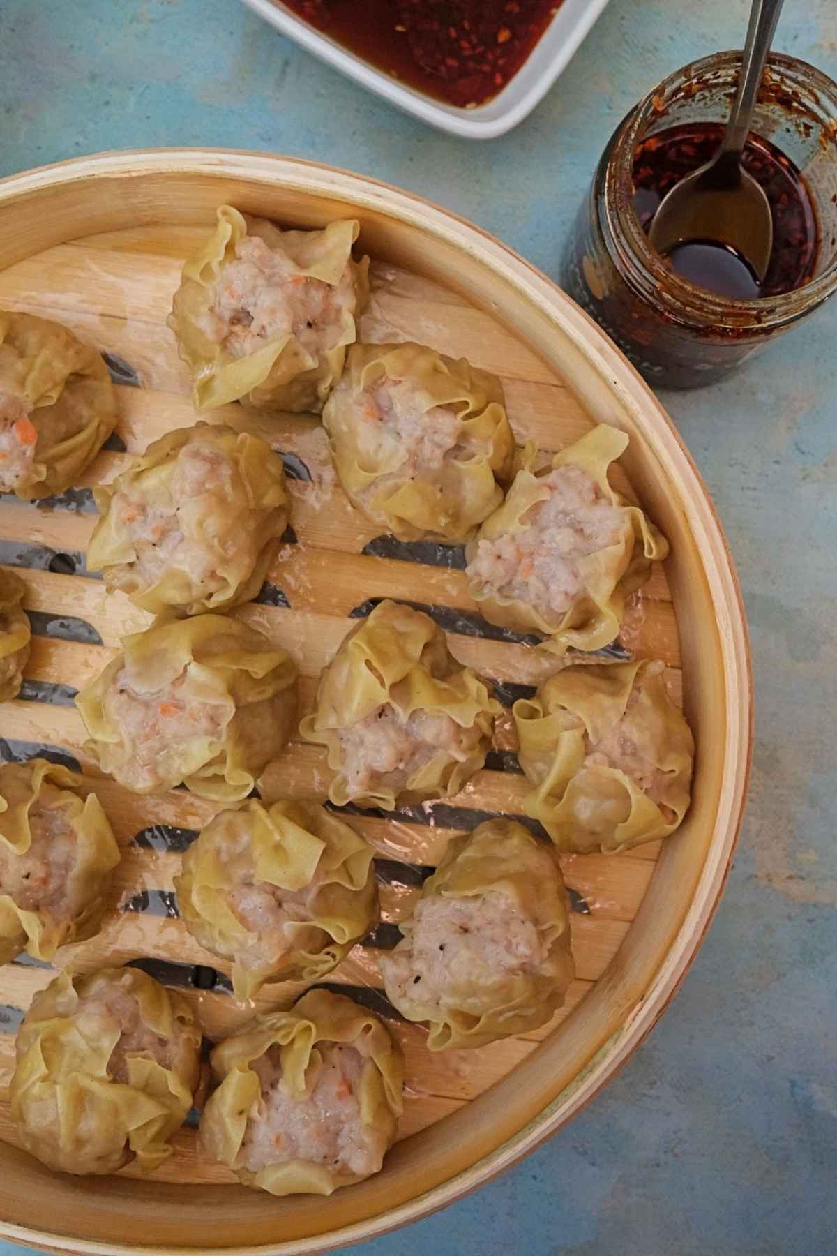 Steamed pork siomai in a bamboo steamer basket served with ponzu sauce and chili garlic on the side.