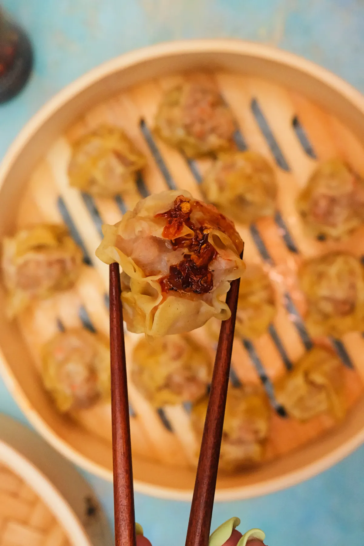 Pork siomai topped with chili garlic held with chopsticks over a bamboo steamer.