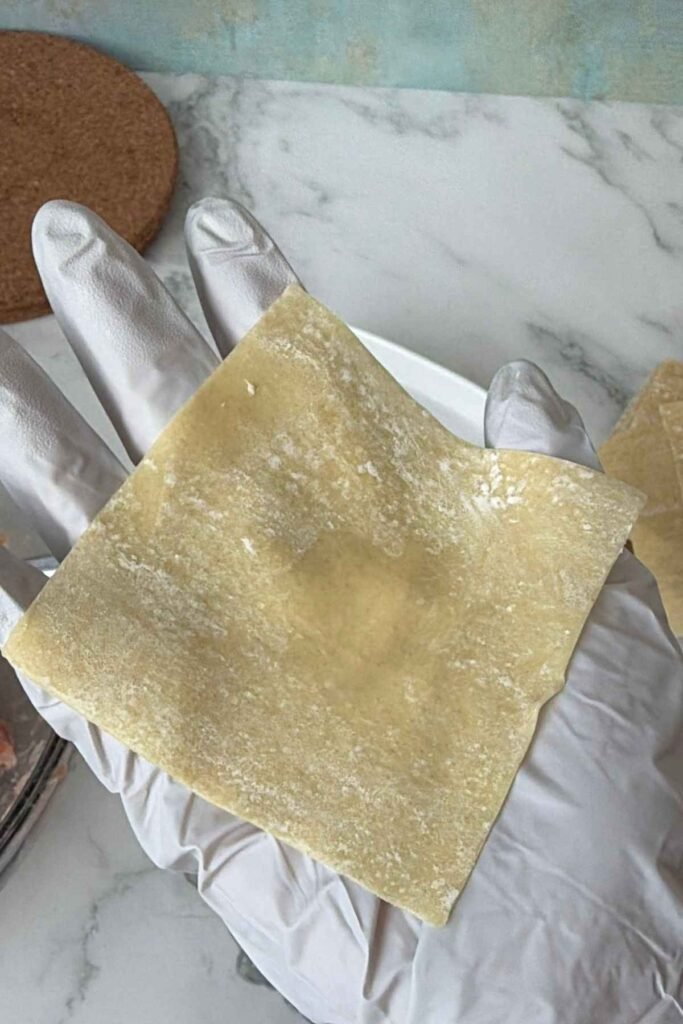 Square extra thin wonton wrapper dusted with flour held in gloved hand.