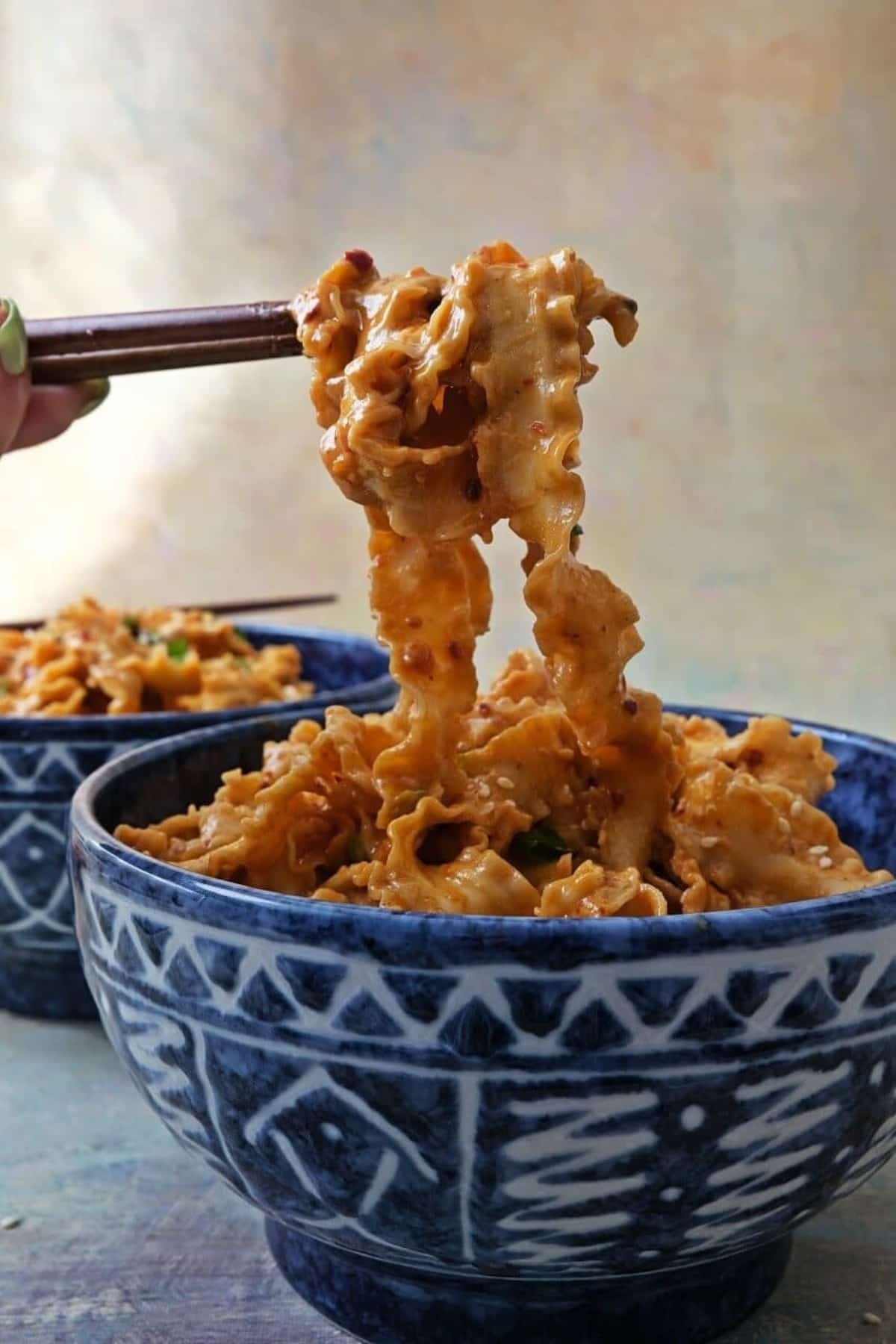 Spicy peanut butter noodles in a bowl, a portion lifted with chopsticks.