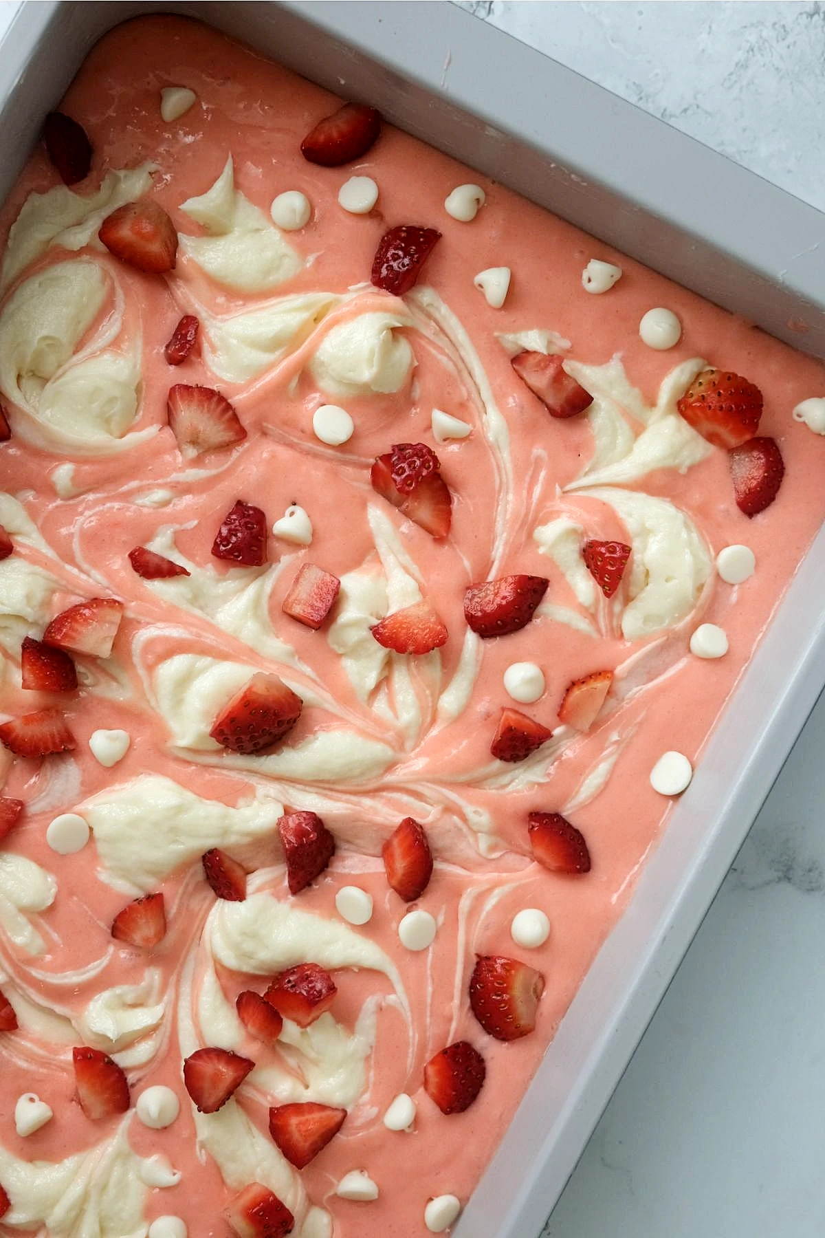 Unbaked strawberry cake batter in a baking dish swirled with cream cheese and topped with diced strawberries and white chocolate chips.