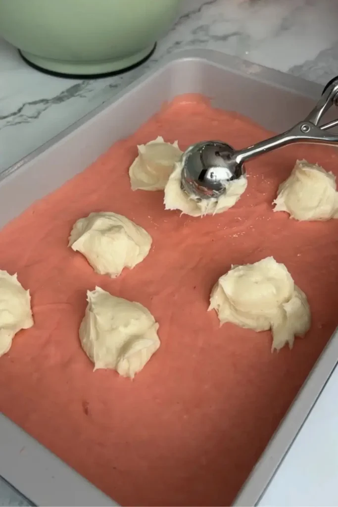 Ice cream scoop dolloping cream cheese mixture evenly over strawberry cake batter in the pan.