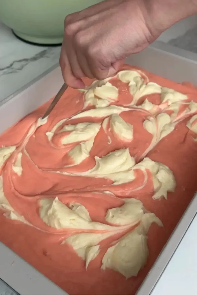 Hand using a knife to swirl cream cheese into strawberry cake batter, creating a marbled pattern.