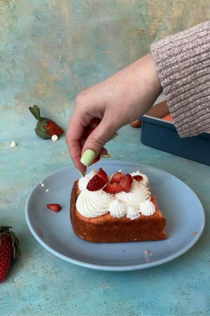 Hand placing sliced strawberries on top of whipped cream frosting over a slice of strawberry earthquake cake on a blue plate.