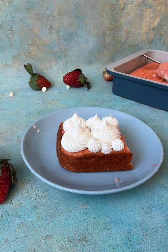 Slice of strawberry earthquake cake topped with piped whipped cream on a blue plate, with fresh strawberries and baking pan in the background.