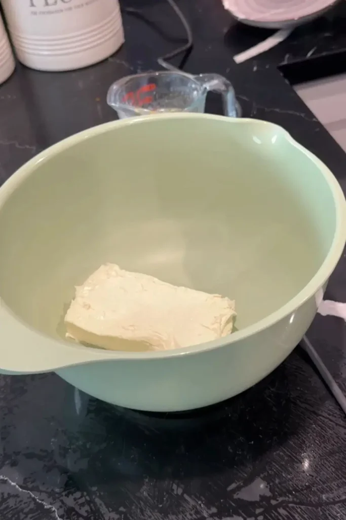 Block of softened cream cheese in a light green mixing bowl on a kitchen counter.