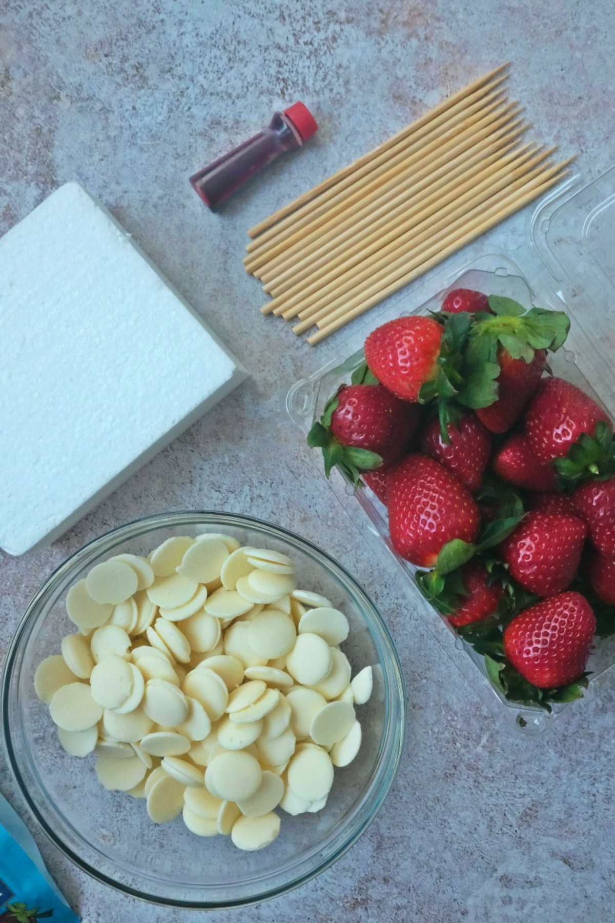 Complete ingredients for white chocolate strawberries: fresh strawberries, white chocolate wafers, red food coloring, wooden skewers, foam block