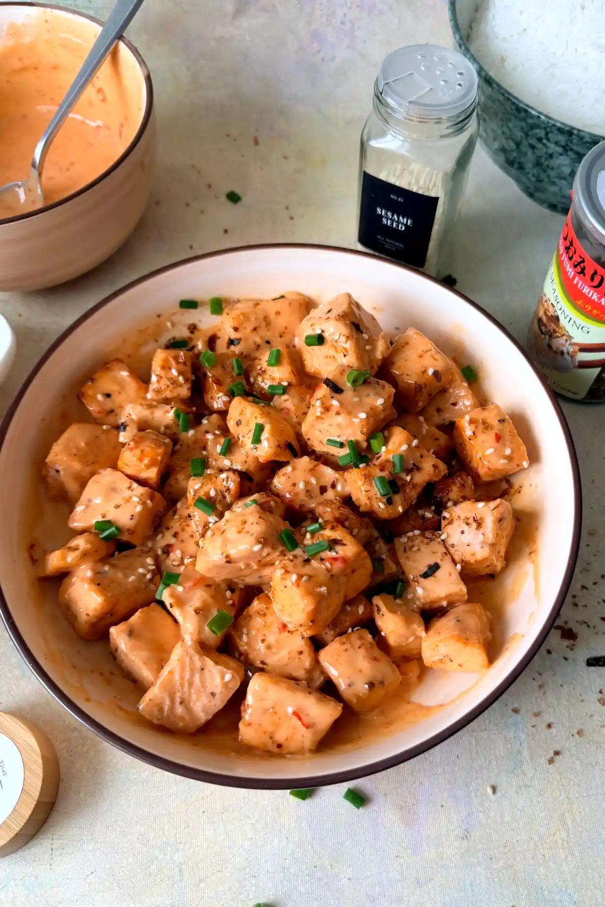 Bowl of saucy bang bang salmon bites garnished with sesame seeds and chives, with sauce and seasonings on the side.