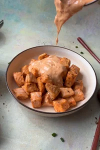 Creamy bang bang sauce being poured over cooked salmon bites in a bowl.