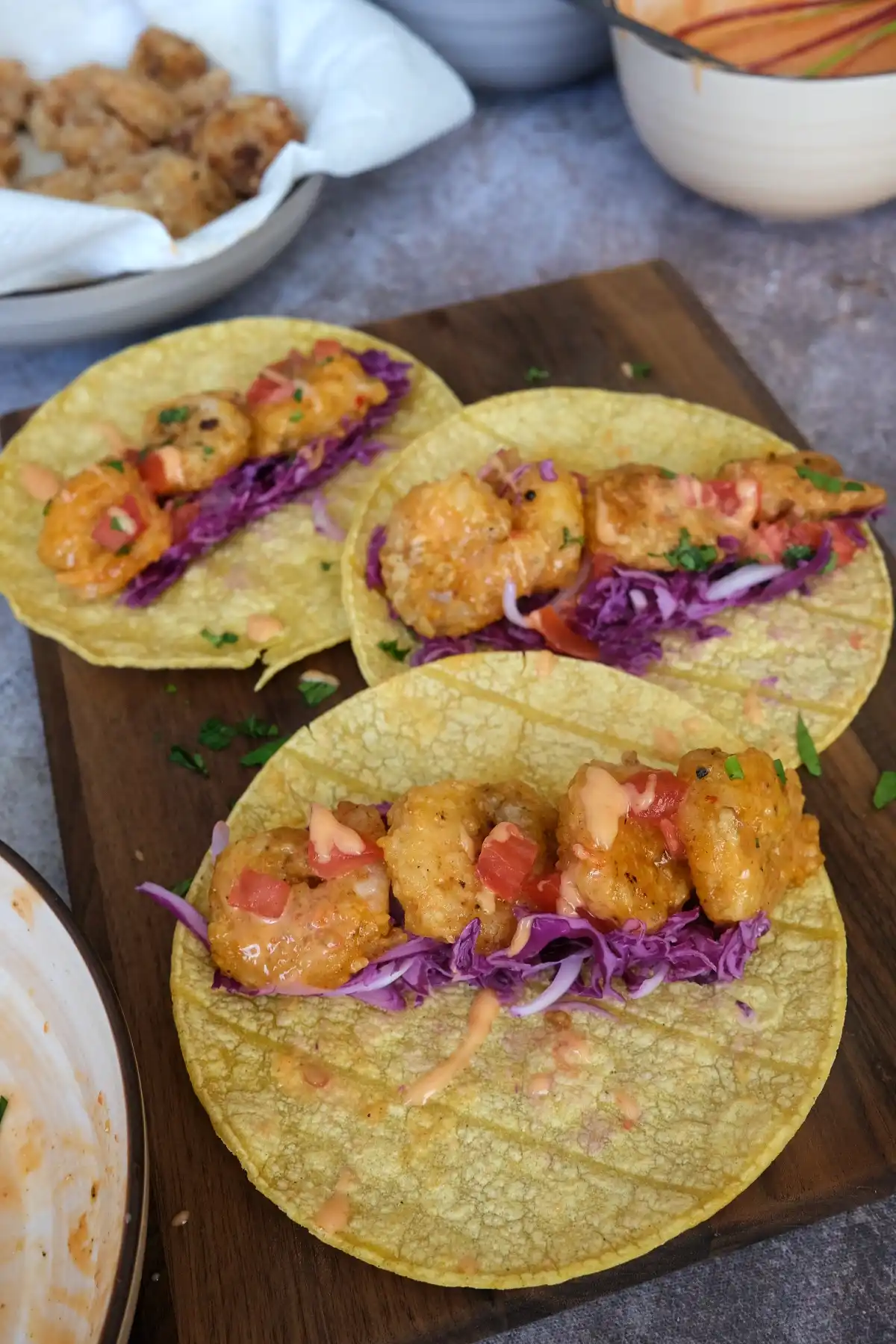 Three bang bang shrimp tacos topped with crispy shrimp, cabbage slaw, tomatoes, and drizzled sauce on a wooden board