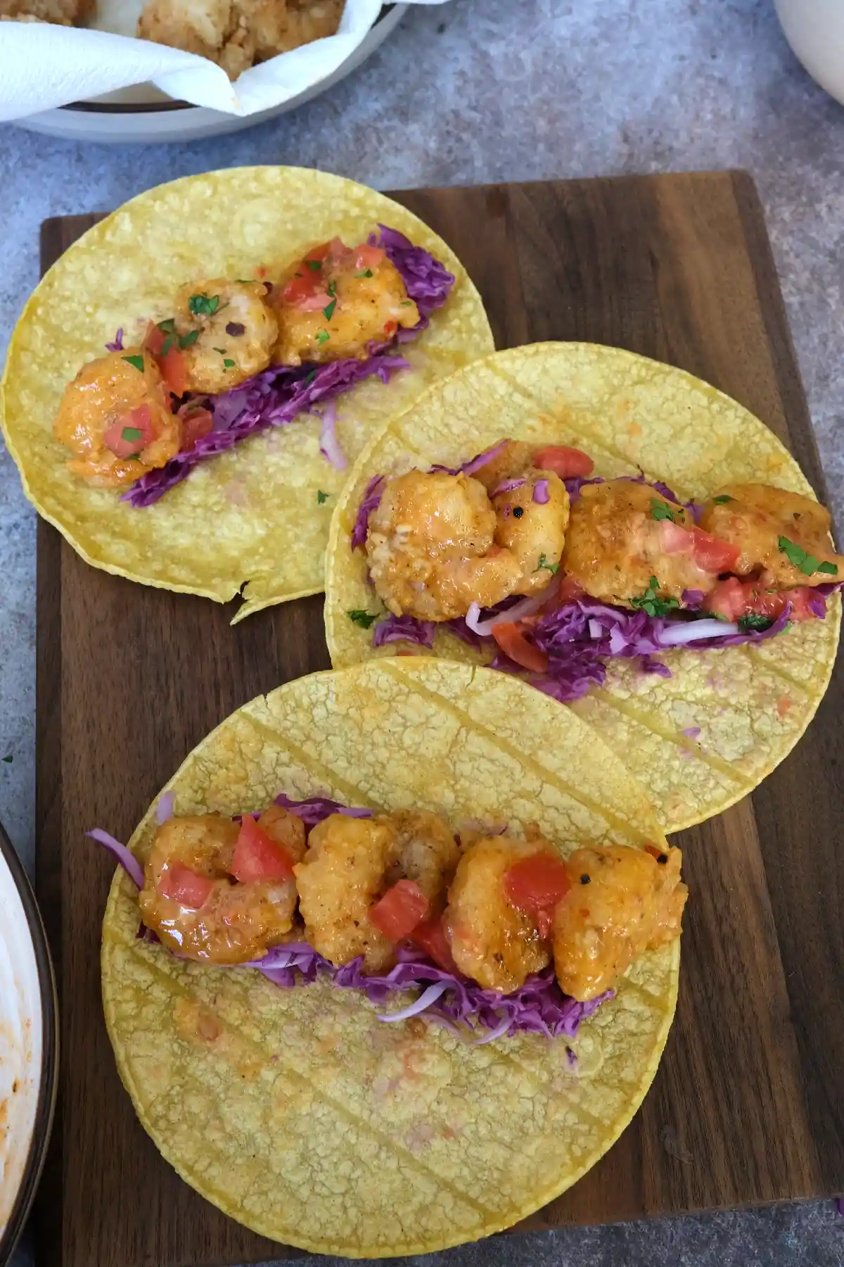 Three bang bang shrimp tacos topped with crispy shrimp, cabbage slaw, tomatoes, and drizzled sauce on a wooden board