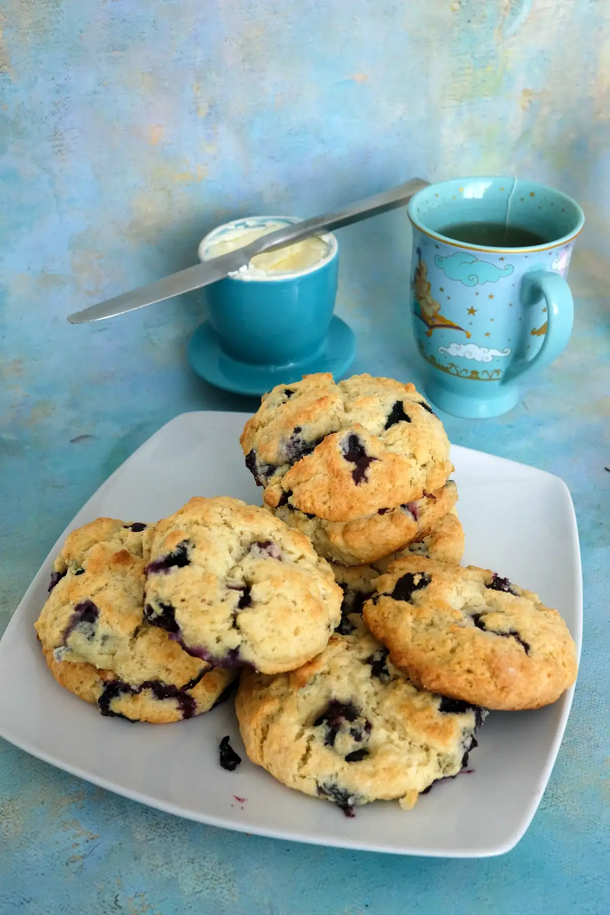 Stack of homemade blueberry biscuits on a plate next to tea and butter.
