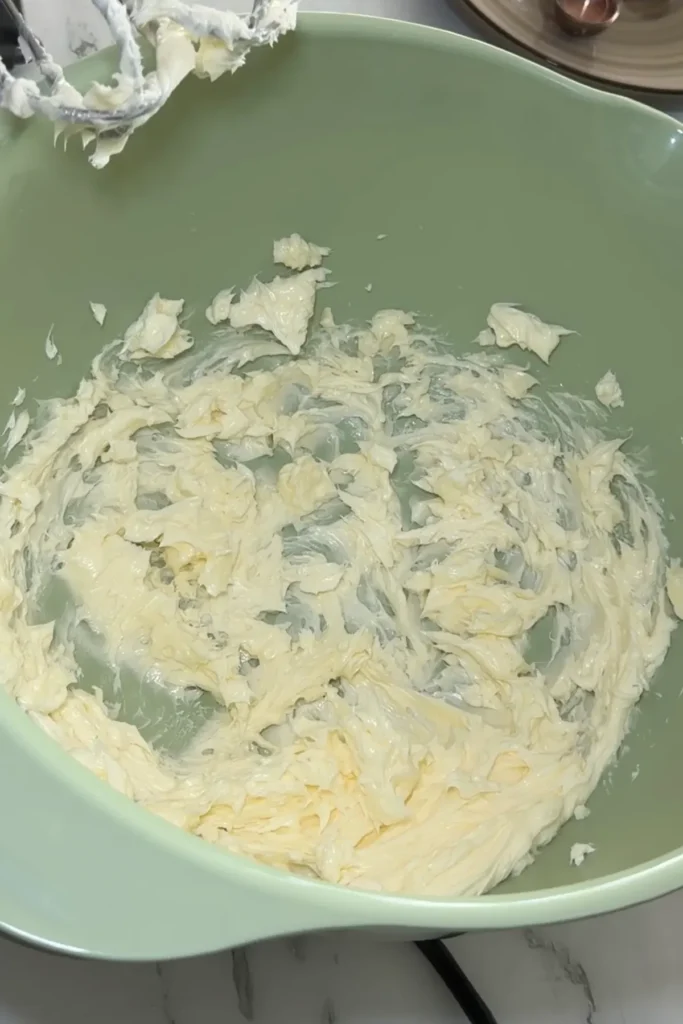 Softened butter creamed in a light green mixing bowl with mixer attachment above.