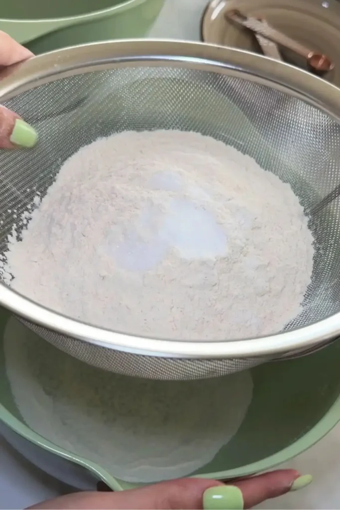 Flour mixture sifted through a fine mesh sieve over a green mixing bowl.