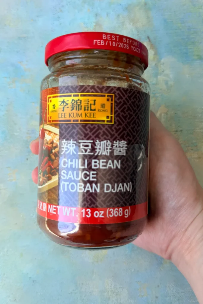 Hand holding a jar of Lee Kum Kee chili bean sauce (toban djan) against a light blue background.