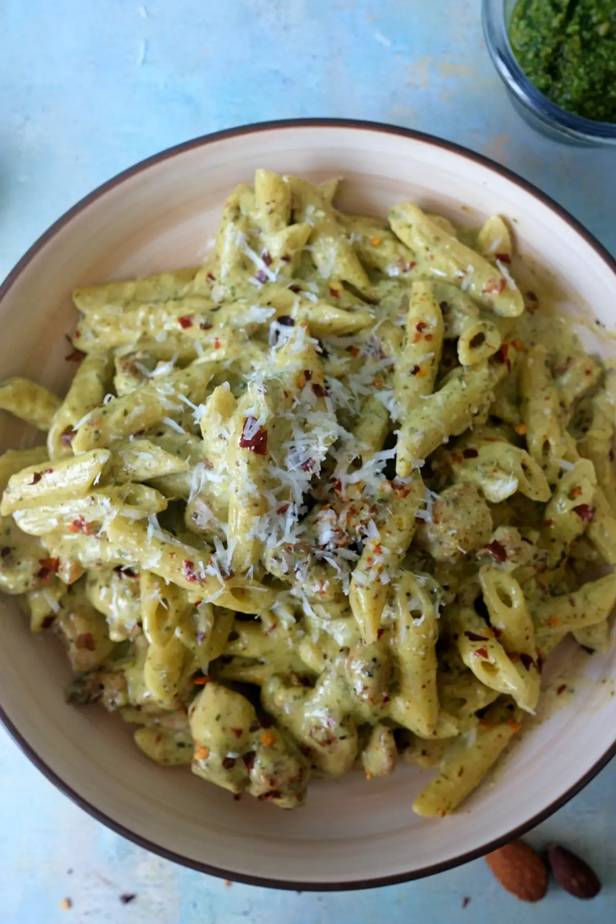 Overhead view of creamy pesto chicken penne pasta topped with parmesan and chili flakes, with a small bowl of pesto partially visible