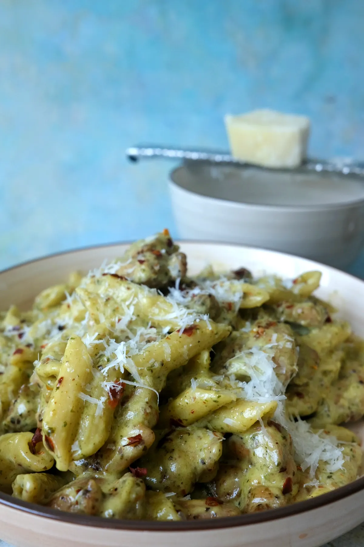 Close-up of creamy pesto chicken pasta with parmesan and chili flakes, with a microplane grater and parmesan block blurred in the background