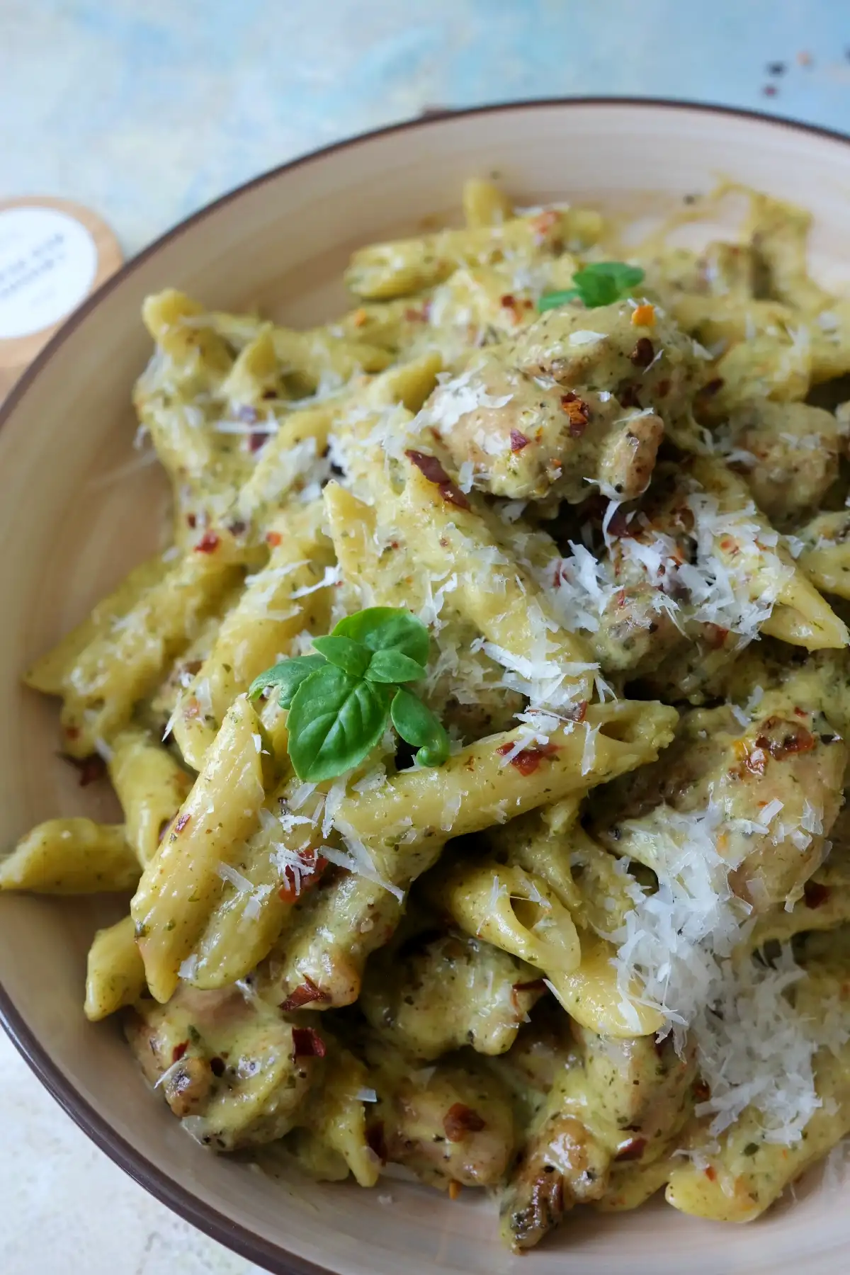 Creamy pesto chicken penne pasta topped with grated parmesan, chili flakes, and a fresh basil leaf in a shallow bowl