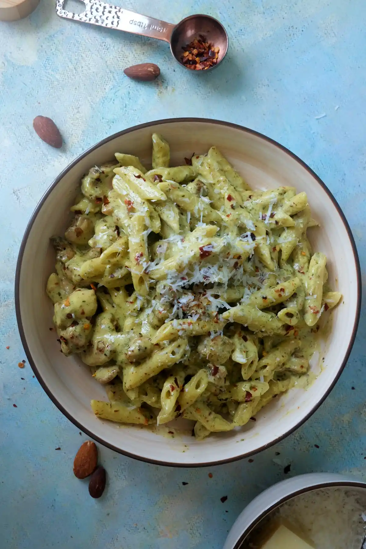 Overhead shot of creamy pesto chicken pasta with parmesan and chili flakes, surrounded by whole almonds and a copper spoon filled with red pepper flakes on a blue surface