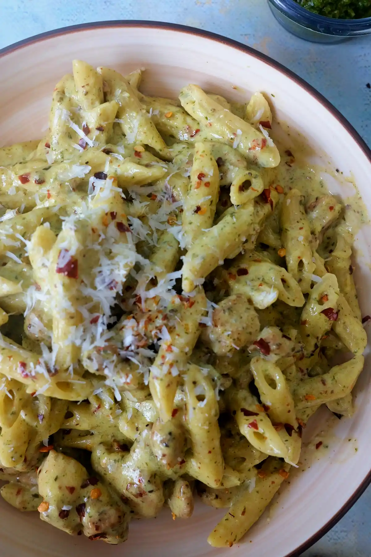 Close-up of creamy pesto chicken penne coated in sauce with parmesan shavings and chili flakes, with a small bowl of pesto in the background