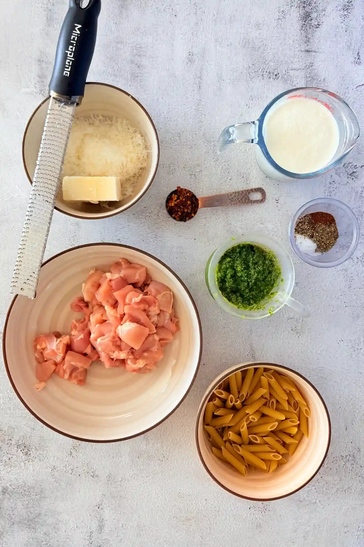 Ingredients for creamy pesto chicken pasta including chicken, penne, pesto, cream, and cheese