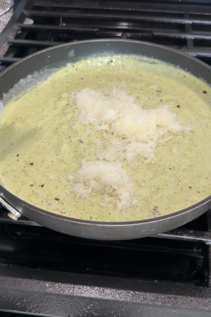 Grated parmesan cheese melting into creamy pesto sauce in skillet