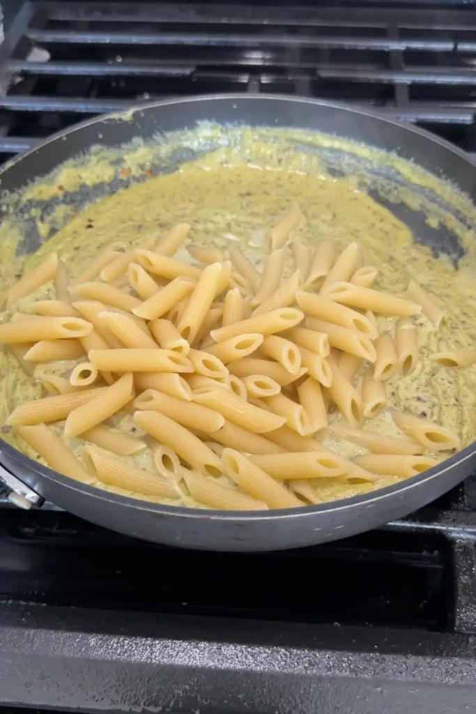 Cooked penne pasta added into creamy pesto sauce in skillet