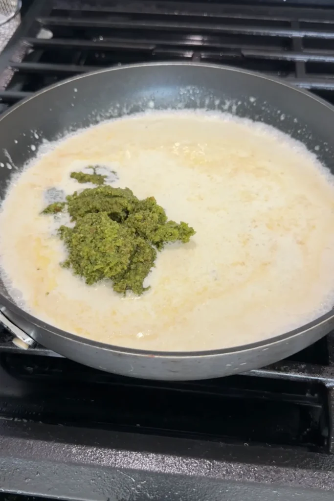 Cream sauce in skillet with a spoonful of green pesto being added