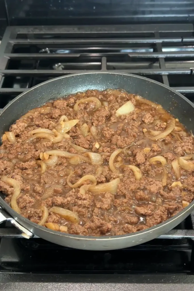 Ground beef and onions being stirred in sauce as it simmers and thickens.