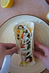 Hands folding a shawarma wrap filled with ground beef, melted cheese, fresh vegetables, and yogurt sauce on a plate