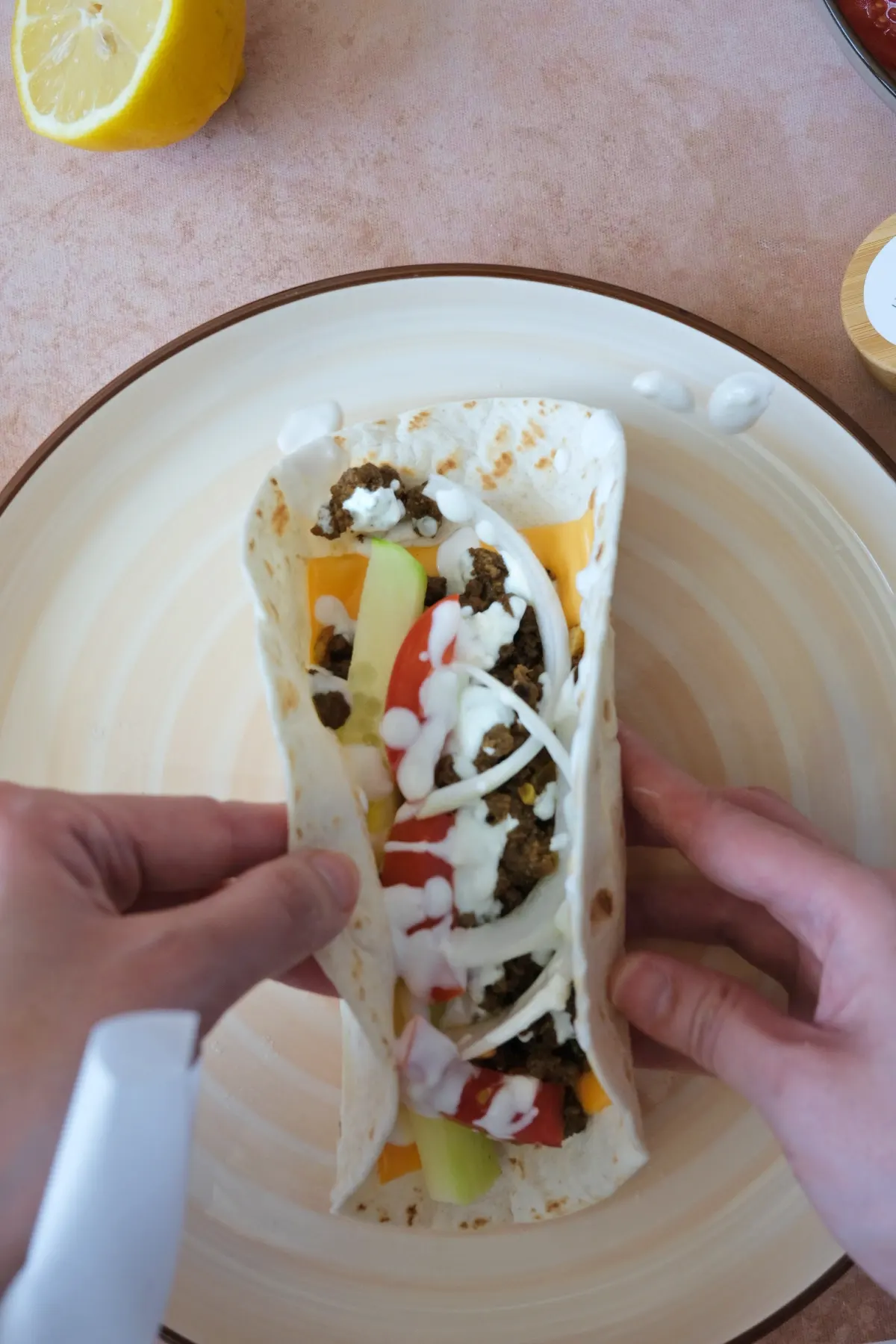 Hands folding a shawarma wrap filled with ground beef, melted cheese, fresh vegetables, and yogurt sauce on a plate