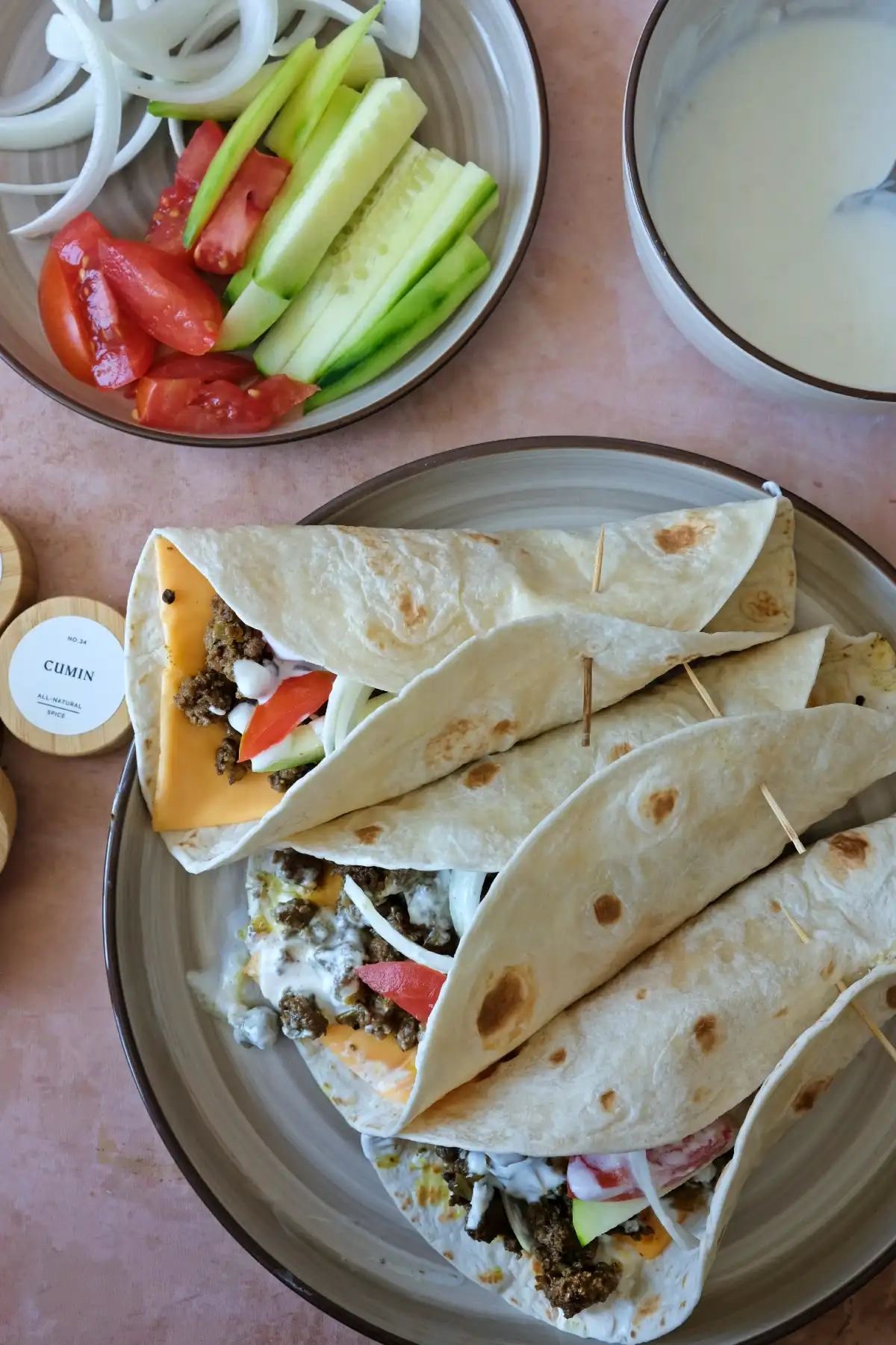Top view of three ground beef shawarma wraps in tortillas with melted cheese, yogurt sauce, and fresh vegetables tucked inside
