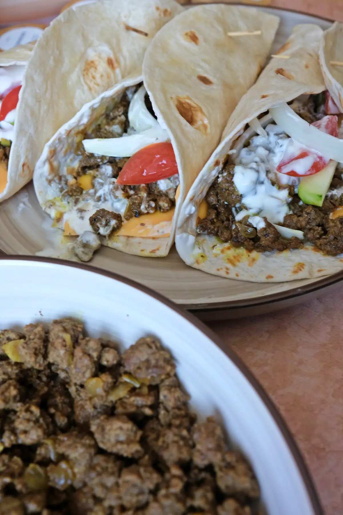 Plate of ground beef shawarma wraps with melted cheese and yogurt sauce alongside a bowl of cooked seasoned beef and fresh sliced vegetables