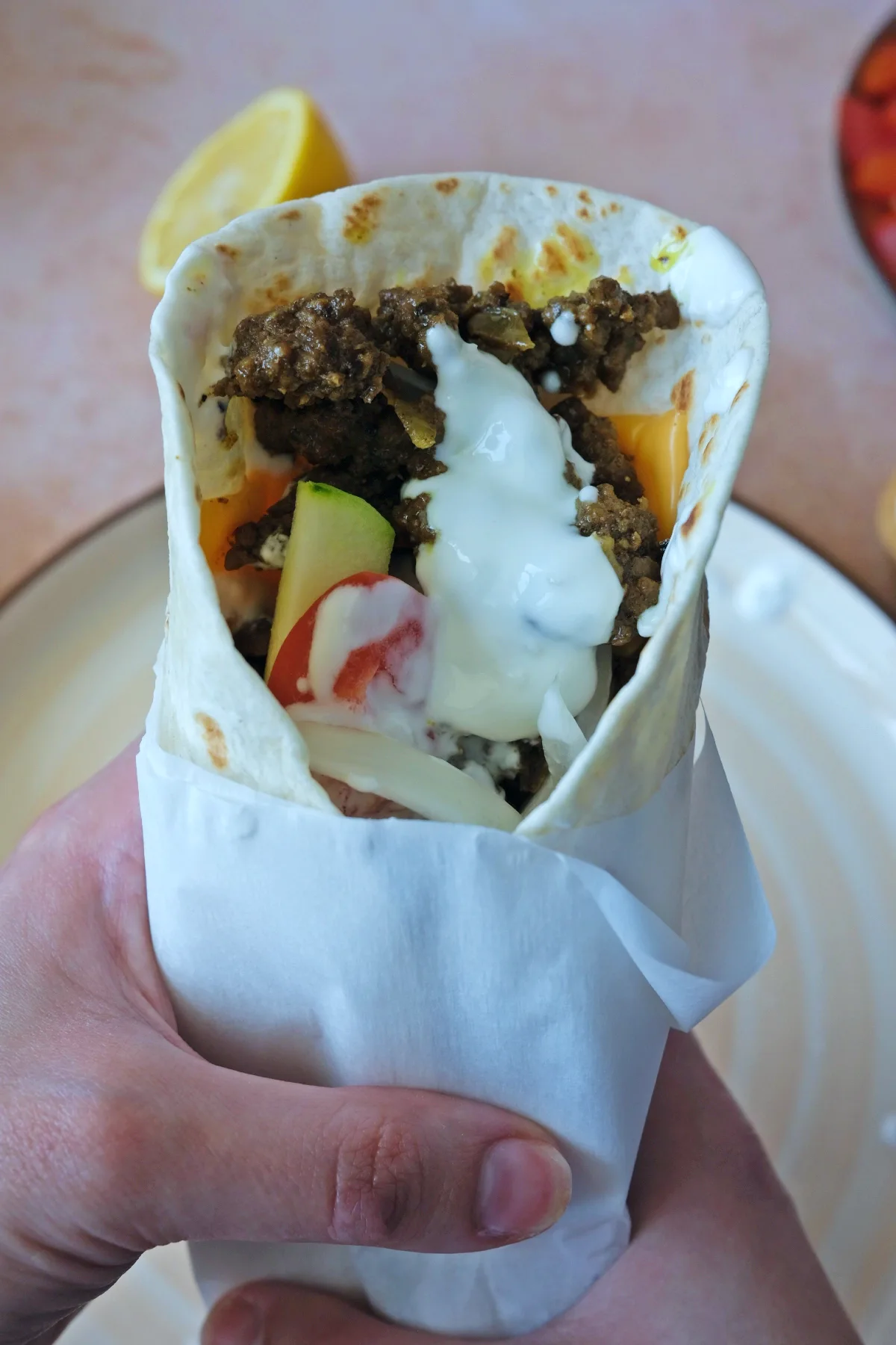 Hand holding a parchment-wrapped ground beef shawarma filled with melty cheese, seasoned beef, vegetables, and yogurt sauce