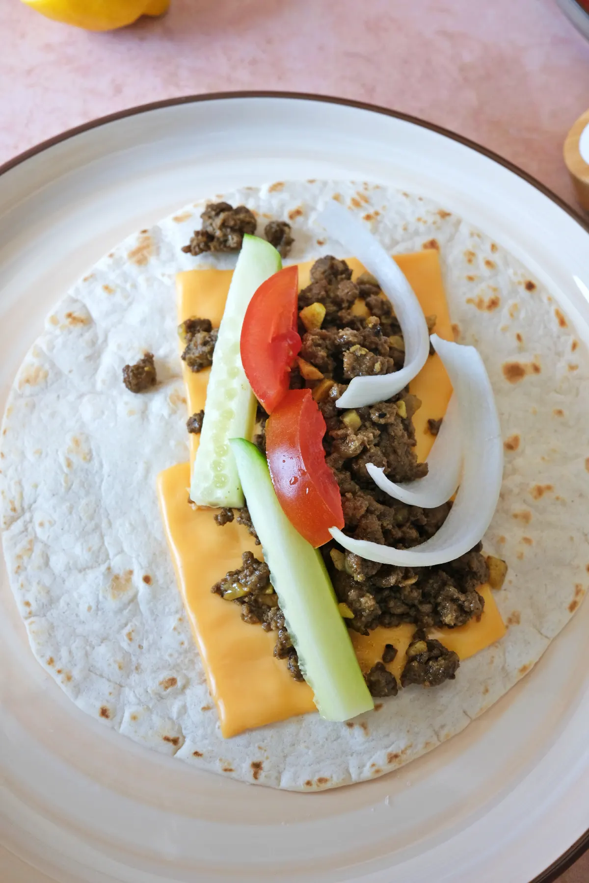 Open flour tortilla with melted American cheese, ground beef shawarma, cucumber sticks, tomato slices, and white onion strips layered before wrapping
